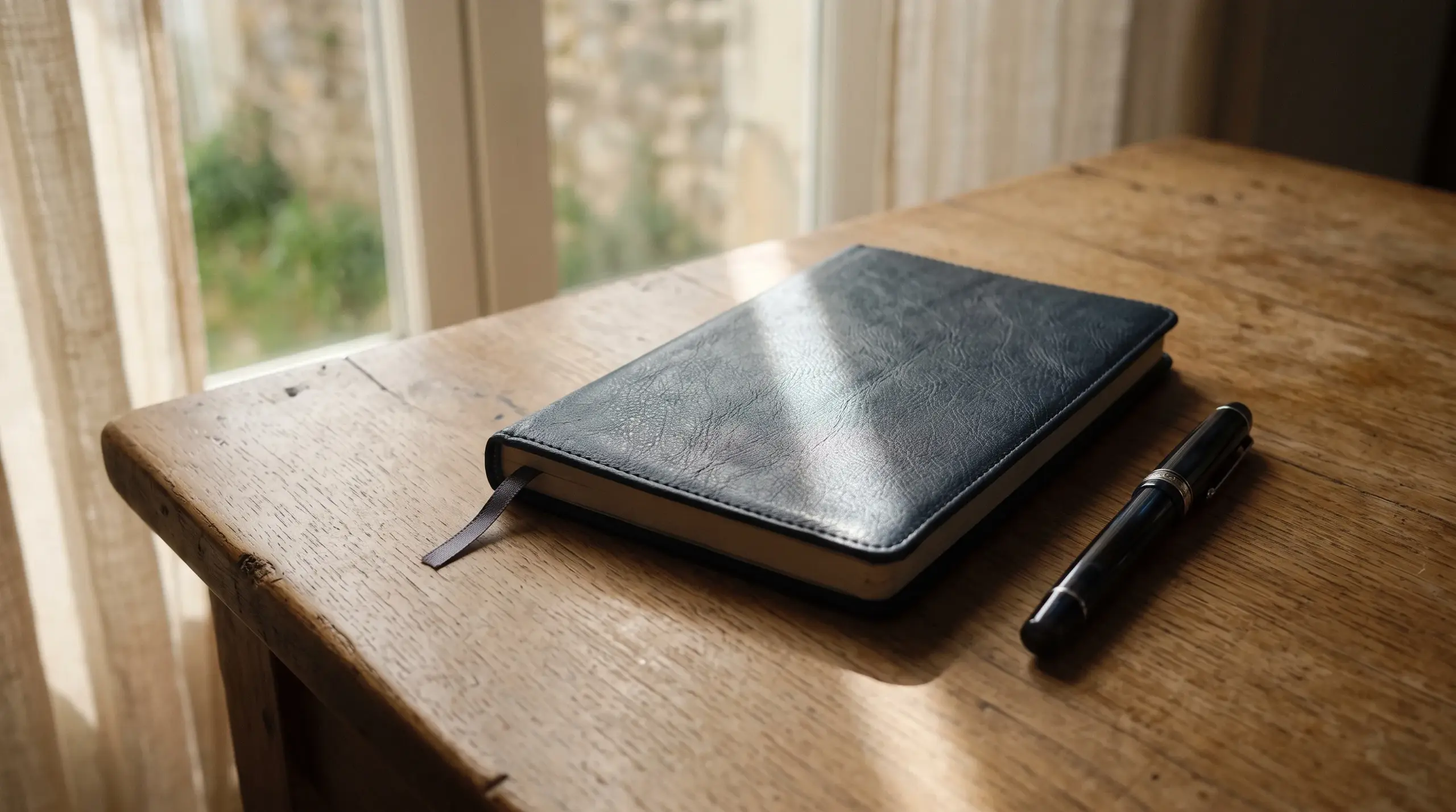 Un carnet fermé et un stylo plume sur une table nue, traversés par un rai de lumière.